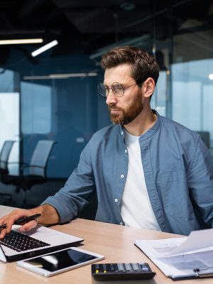 A serious young man accountant, financier, analyst, auditor sits in the office at the table. He holds documents and a pen in his hands, checks accounts, finances, types on a laptop.