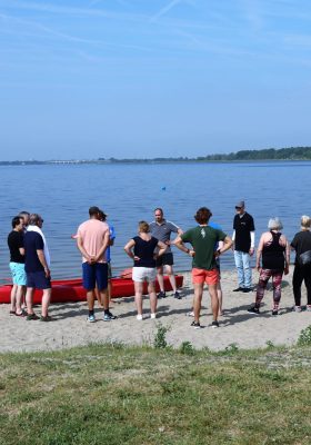 Een groep mensen op het strand die naar een rode kano kijken. Een foto van het uitje van het ORdigiNAL 2022 team.