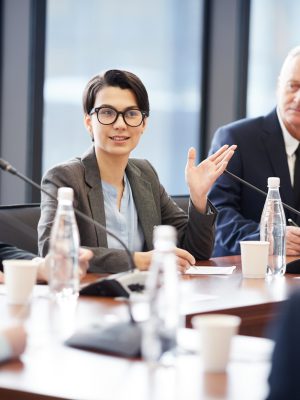 Portrait of young businesswoman speaking to microphone during group discussion in conference room