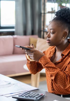 Jonge Afrikaanse vrouwelijke accountant in slimme vrijetijdskleding die foto van financiële documenten op lijst neemt terwijl het werken in huismilieu