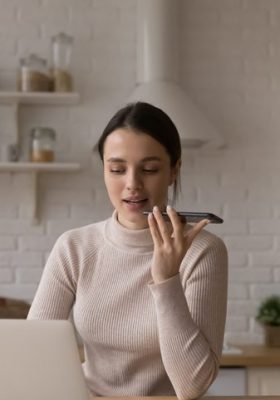 Woman sit in kitchen works on laptop and speaks to phone to dictate.