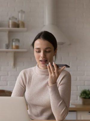 Une femme assise dans la cuisine travaille sur son ordinateur portable et dicte au téléphone.