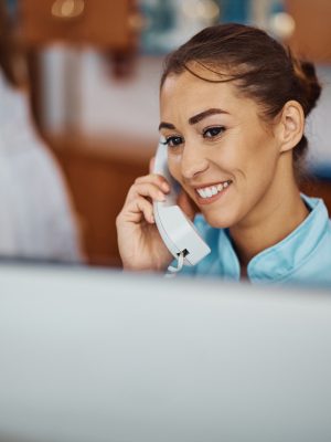 Junge Krankenschwester, die einen Anruf entgegennimmt, während sie an einem Desktop-PC an der Rezeption eines Krankenhauses arbeitet. Platz zum Kopieren.