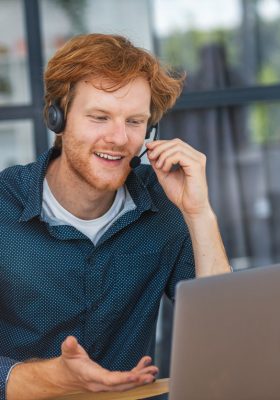 Giovane uomo caucasico, operatore del call center di assistenza clienti, seduto sul posto di lavoro in un ufficio moderno che consulta un cliente, utilizza una cuffia, sorride amichevolmente. Viene utilizzata la telefonia