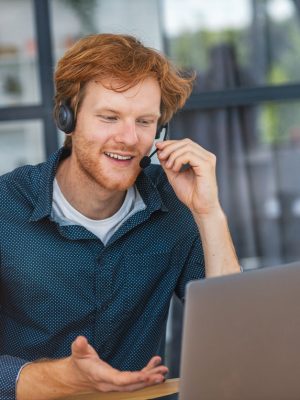 Giovane uomo caucasico, operatore del call center di assistenza clienti, seduto sul posto di lavoro in un ufficio moderno che consulta un cliente, utilizza una cuffia, sorride amichevolmente. Viene utilizzata la telefonia