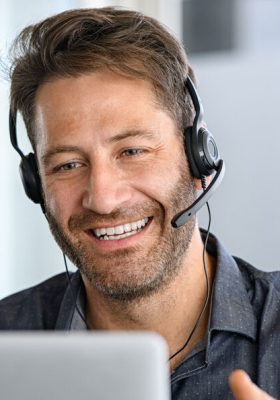 Technical support operator working with headset in office. Smiling handsome man working as call centre operator, speaking to customer and using laptop. Happy successful businessman working remotely while doing video conference.