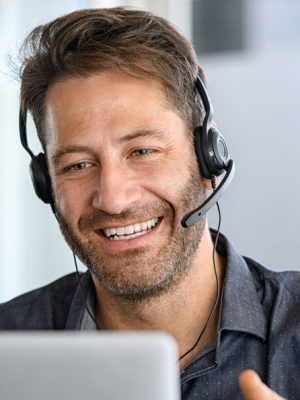 Technical support operator working with headset in office. Smiling handsome man working as call centre operator, speaking to customer and using laptop. Happy successful businessman working remotely while doing video conference.