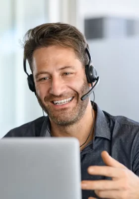 Technical support operator working with headset in office. Smiling handsome man working as call centre operator, speaking to customer and using laptop. Happy successful businessman working remotely while doing video conference.