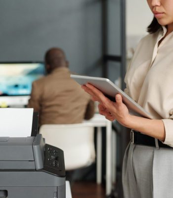 Focus on tablet held by businesswoman computing in office while standing by printer. Powered by Document Automation Solutions