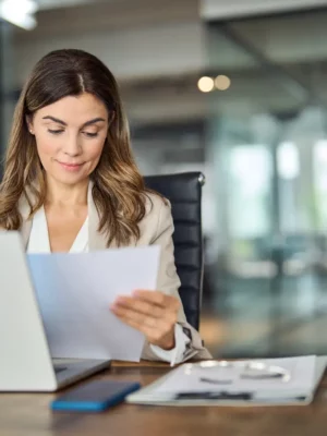 Busy mittleren Alters Business-Frau arbeitet im Büro mit Laptop lesen Dokument. Mature professionellen weiblichen Manager Anwalt Anwalt hält Papier Finanzbericht sitzt am Schreibtisch im Büro. Kopieren Raum