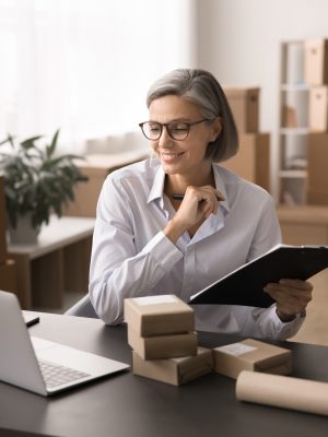 Smiling professional woman, small business owner or warehouse worker looks at laptop screen, holds clipboard and pen, parcels stacked at desk, involved in office work, managing orders or logistic task