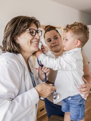 Smiling pediatrician lets a young boy use her stethoscope while his mother watches during a check-up.