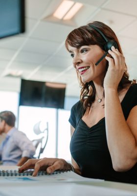 Mooie rijpe zakenvrouw die op headset praat terwijl ze aan haar bureau werkt. Kaukasische vrouwelijke leidinggevende met mensen die op achtergrond werken.