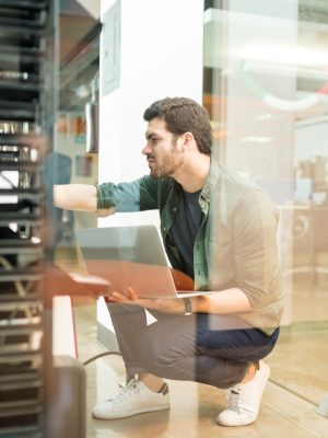 Network administrator with notebook computer sitting in cloud center room and working with networking device on rack cabinet