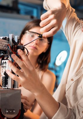 Junger Mann und Frau mit Schutzbrille bei Experimenten in der Robotik in einem Labor. Roboter auf dem Tisch