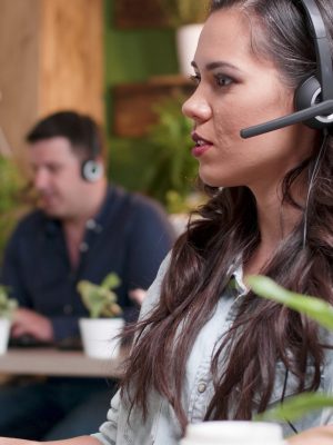 Junge Kundenbetreuerin, die in ein Headset-Mikrofon mit einem Callcenter-Kunden spricht, während sie im Büro eines Startup-Unternehmens arbeitet. Kaukasische Beraterin, die Telemarketing-Dienstleistungen anbietet